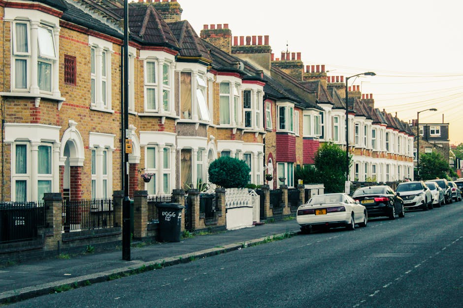 View of a residential street in Highbury, Islington, showing a row of Victorian-style brick houses with white decorative trim and black iron fences along the pavement. In the foreground, a blooming tr