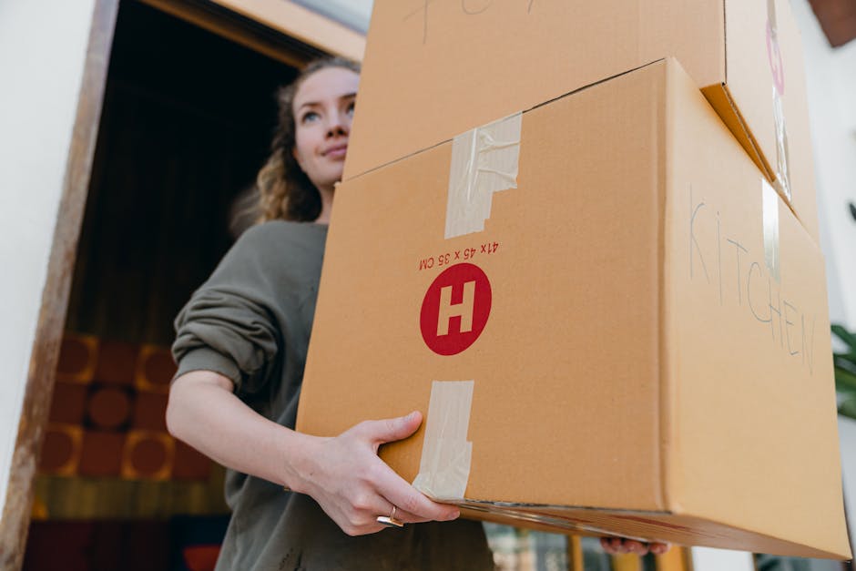 A woman with curly hair, dressed in a grey long-sleeve top, is seen holding a large cardboard box during a home relocation process. She is standing inside a building, near a doorway, with wooden furni
