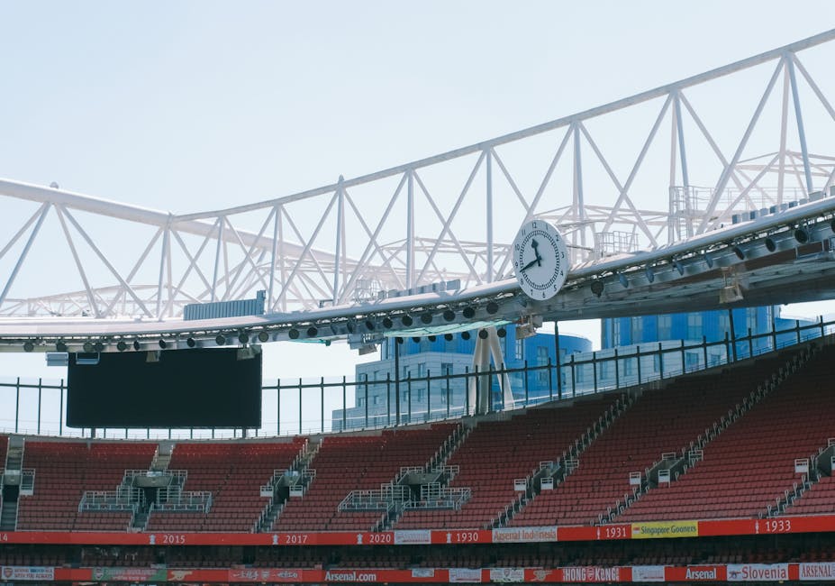 Within a sports stadium, a section of the seating area with red chairs is visible, along with a black digital screen mounted on a metal railing. Above, a large white structural framework supporting th