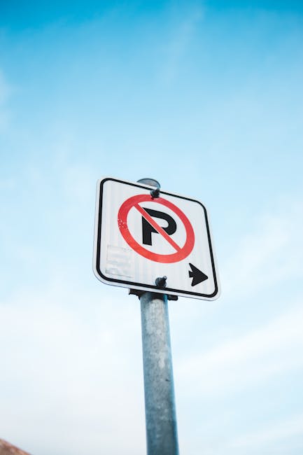 A vertically mounted rectangular metal sign with a white background and red border affixed to a corrugated metal wall. The sign displays the message 'DON'T EVEN THINK OF PARKING HERE' in bold red lett